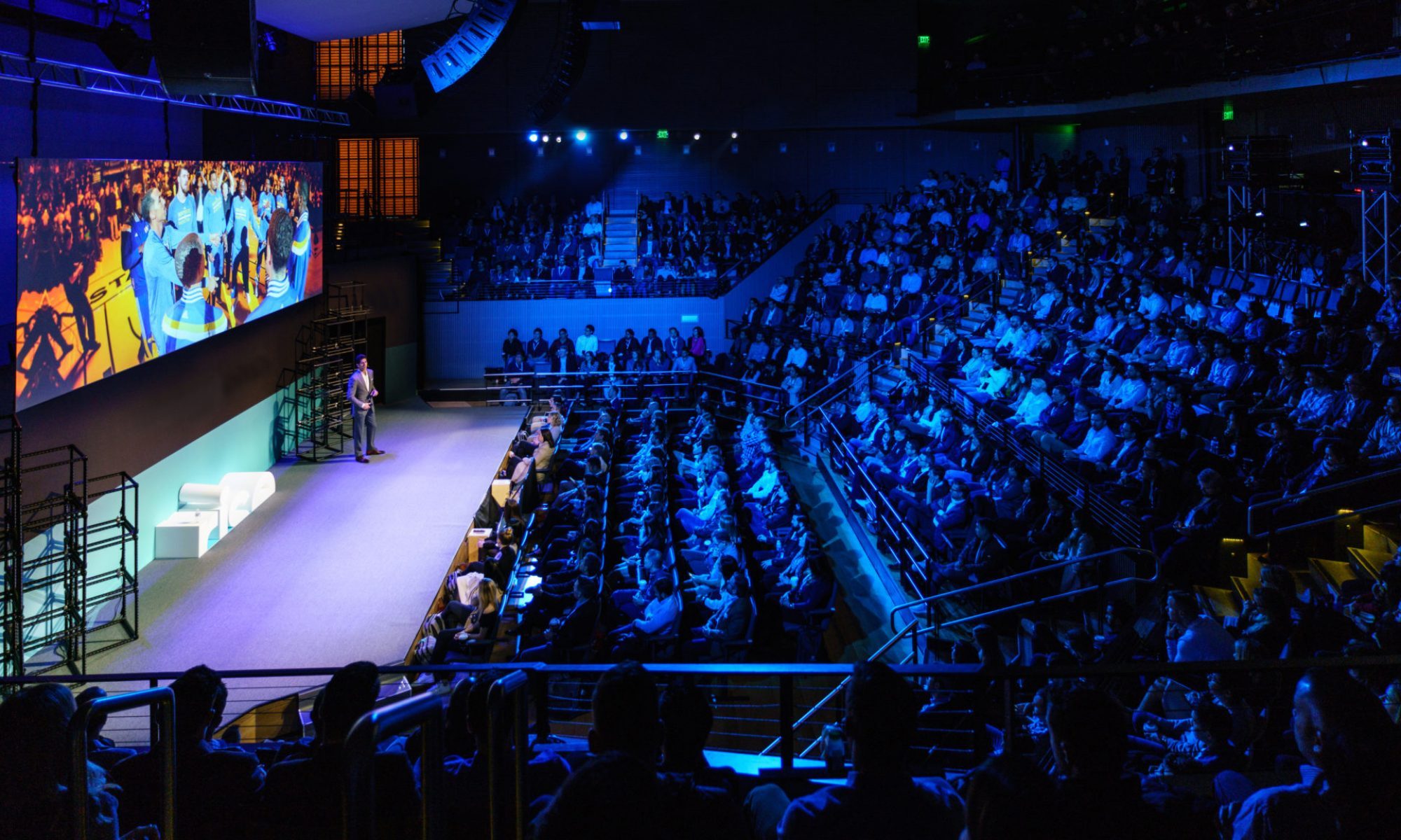 A packed auditorium lit in blue, with a speaker on stage beside a large LED screen